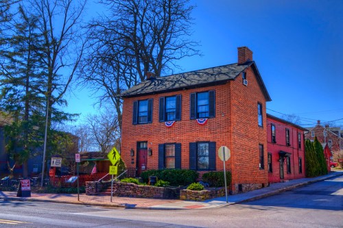 A brick house with black shutters, surrounded by trees, under a clear blue sky, featuring patriotic decorations.