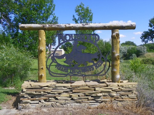 A rustic wooden sign with a metal cutout of a horse, surrounded by greenery and blue sky.