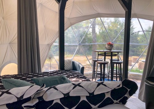 Cozy interior of a geodesic dome with a bed, small dining table, and large windows overlooking nature.
