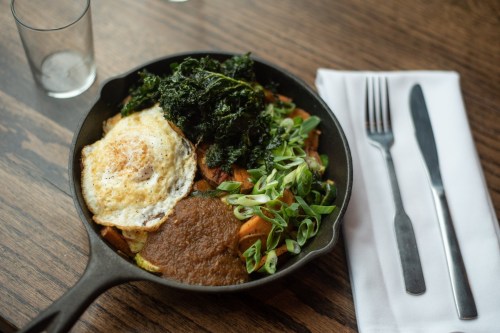 A cast iron skillet filled with sautéed vegetables, a fried egg, and greens, served on a wooden table with cutlery.