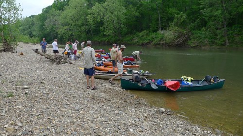A group of people preparing canoes by a riverbank surrounded by greenery on a cloudy day.