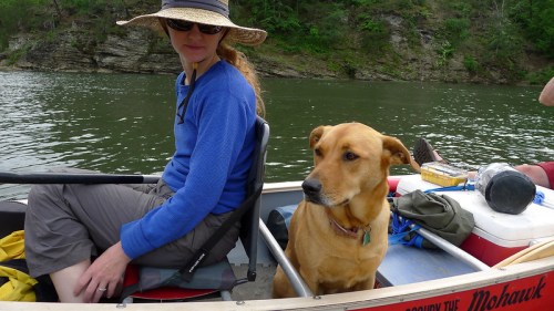 A woman in a hat sits in a boat beside a dog, with a scenic river and greenery in the background.