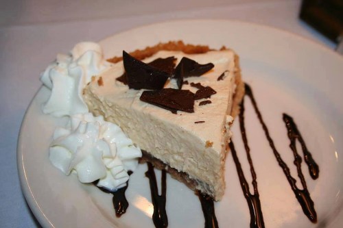 A slice of creamy cheesecake topped with chocolate shavings, served with whipped cream and chocolate drizzle on a plate.