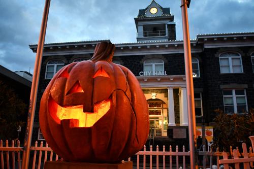 A large carved pumpkin with a smiling face in front of a historic city hall building at dusk.