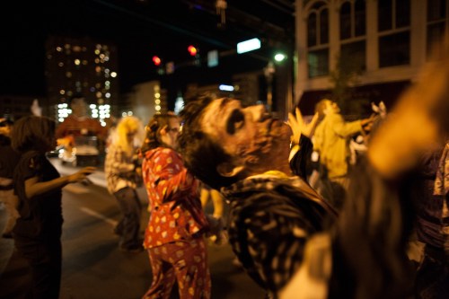 A crowd of people in costumes dancing on a city street at night, with buildings and traffic lights in the background.