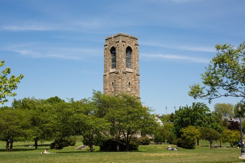 A stone tower stands in a park surrounded by green trees and a clear blue sky, with people relaxing on the grass.