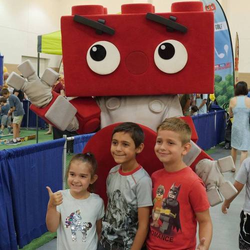 A large, friendly mascot poses with three smiling children at a lively event, surrounded by colorful banners.