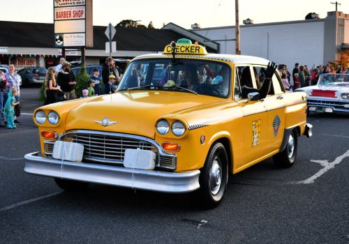 A classic yellow taxi cab with a checker logo drives through a parade, surrounded by onlookers and festive decorations.