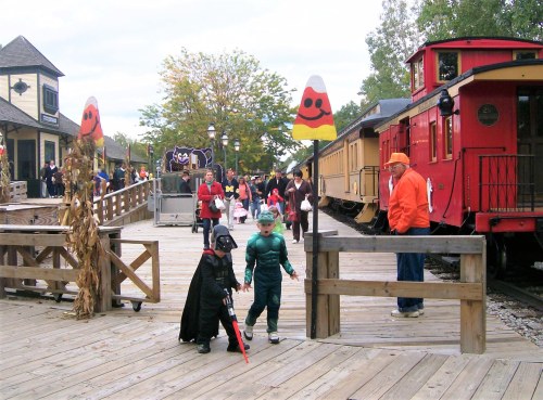 Children in costumes walk along a wooden path near colorful train cars, with festive decorations for Halloween.