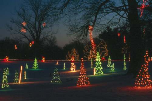 Colorful Christmas lights illuminate a snowy landscape, featuring decorated trees and festive figures against a twilight sky.