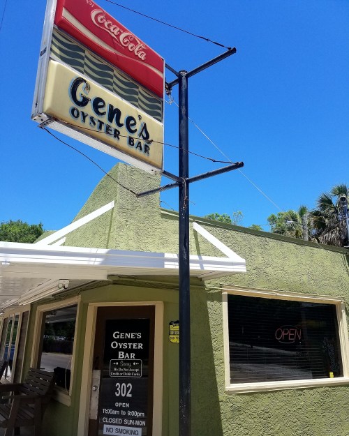 Sign for Gene's Oyster Bar with a Coca-Cola logo, featuring an open sign and a green exterior under a clear blue sky.