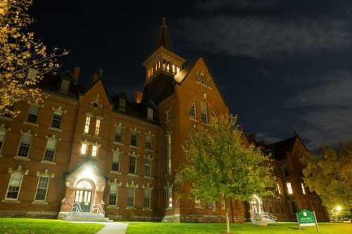 Historic brick building illuminated at night, featuring a tall spire and surrounding trees under a cloudy sky.