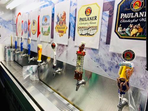 A row of beer taps with various colorful handles, featuring Paulaner beer labels in a festive setting.