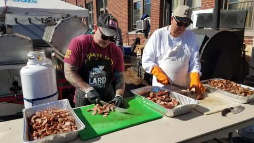 Two men prepare and chop sausage on a table outdoors, with cooking equipment and a building in the background.