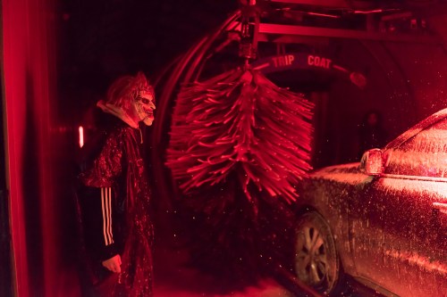 A person in a mask stands beside a car in a car wash, illuminated by red lights and surrounded by swirling soap.