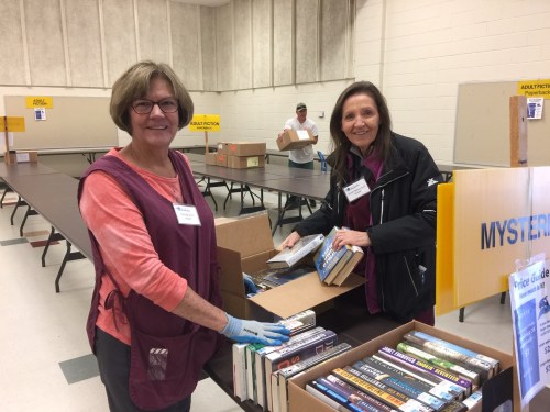 Two women sort books at a community event, with tables labeled for different genres in the background.