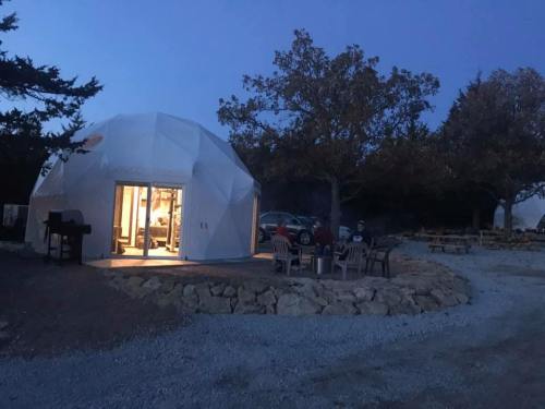 A geodesic dome structure at dusk, surrounded by trees and gravel, with outdoor seating and a grill nearby.