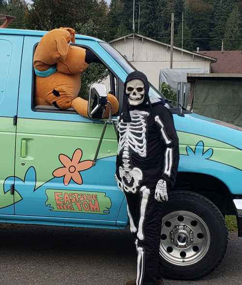 A person in a skeleton costume stands next to a colorful van with a large Scooby-Doo character inside.