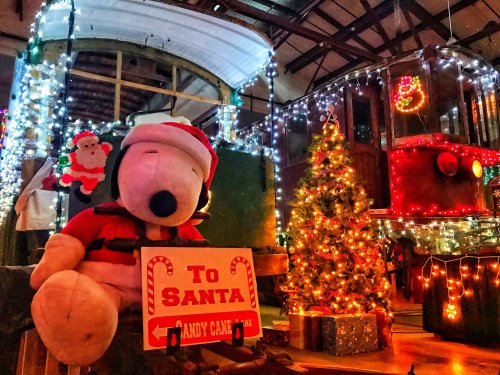 A festive scene featuring a large stuffed Snoopy in a Santa hat, surrounded by Christmas lights and a decorated tree.