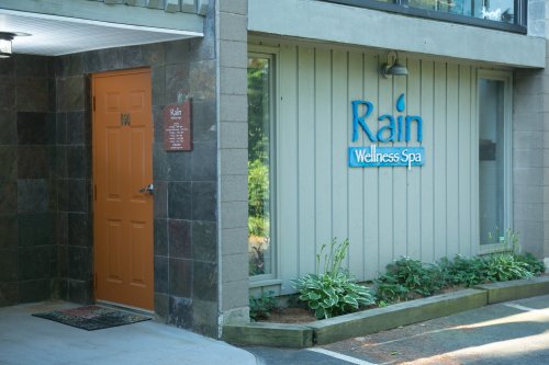Exterior view of Rain Wellness Spa, featuring an orange door and a sign with the spa's name. Lush greenery surrounds the entrance.