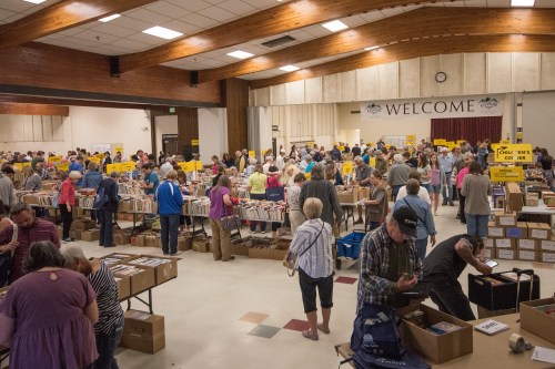 A large crowd browses tables filled with boxes of books in a spacious indoor venue. Signs indicate various sections.