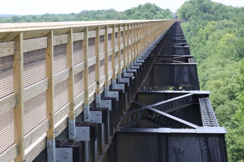 A wooden railing along a high metal bridge, overlooking a lush green landscape below.