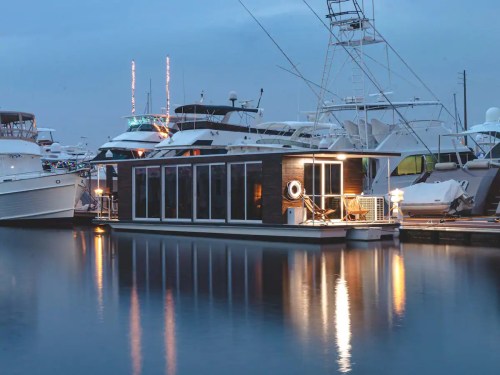 A modern houseboat illuminated at dusk, docked among larger yachts in a marina. Reflections shimmer on the water.
