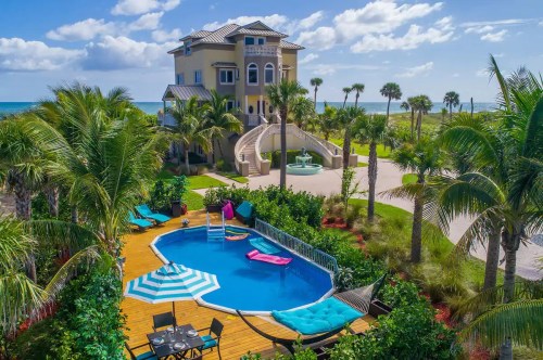 A luxurious beachfront home with a pool, surrounded by palm trees and a sunny sky, featuring a slide and outdoor seating.