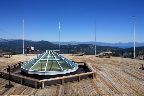 View from a wooden observation deck featuring a glass pyramid structure, with mountains and a clear blue sky in the background.