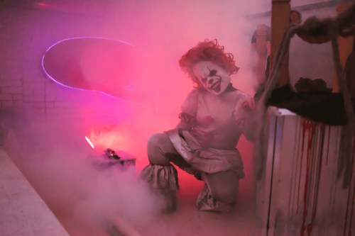A creepy clown in a foggy, dimly lit space, surrounded by eerie lighting and a sinister atmosphere.
