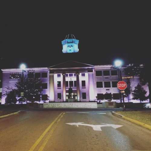 Historic building illuminated at night, featuring a clock tower and purple lighting, with a stop sign in the foreground.