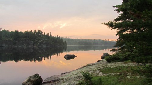 Serene lake at sunrise, surrounded by trees and rocky shores, reflecting soft pastel colors in calm water.