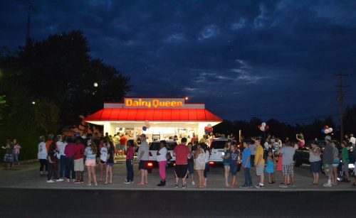 A crowd gathers outside a Dairy Queen under a darkening sky, with balloons and festive decorations visible.