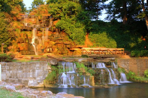 A serene waterfall cascading over rocks, surrounded by lush greenery and a wooden bridge in a tranquil park setting.