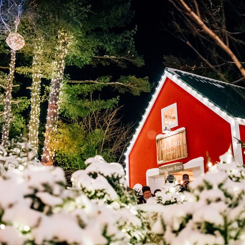 A cozy red barn surrounded by snow and festive lights, with trees and decorations creating a winter wonderland atmosphere.