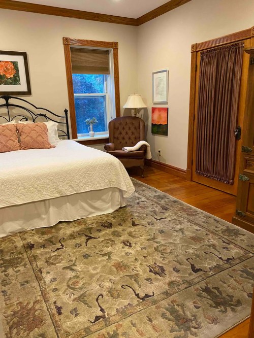 Cozy bedroom with a bed, armchair, lamp, and decorative rug, featuring warm wood accents and soft lighting.