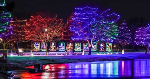 Colorful holiday lights illuminate trees and displays by a calm lake at night, creating a festive atmosphere.