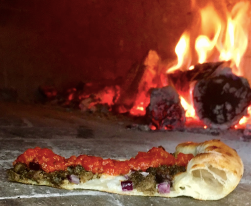 A slice of pizza topped with sauce and toppings, resting on a stone surface near a glowing wood-fired oven.
