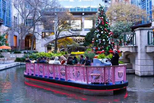A festive boat with passengers cruises along a decorated canal, featuring a Christmas tree and holiday decorations.