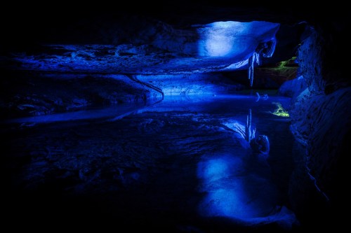 A dark cave with blue lighting reflecting on still water, showcasing unique rock formations and icicles.