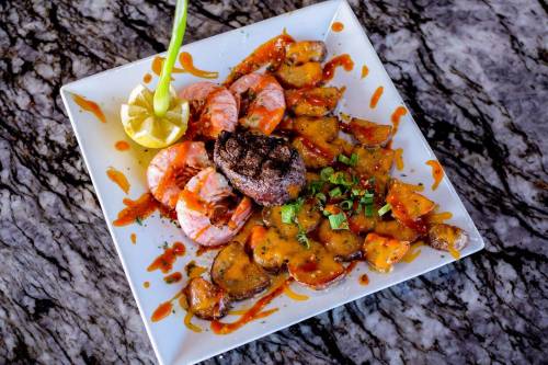 A plate of grilled shrimp and steak with a spicy sauce, garnished with green onions and a lemon wedge.