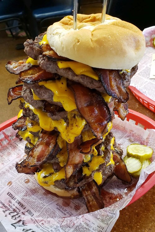 A towering burger stacked with multiple beef patties, melted cheese, and crispy bacon, served in a red basket.