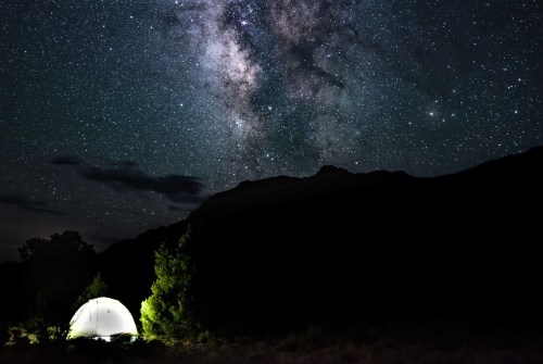 A tent illuminated by a soft light under a starry night sky filled with the Milky Way and distant mountains.