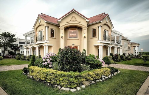 A large, elegant house with a landscaped garden, featuring flowers and greenery, under a cloudy sky.