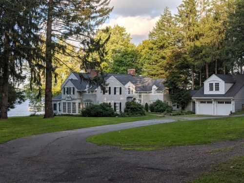 A large, elegant house surrounded by trees, with a driveway leading to a garage and a view of a lake in the background.