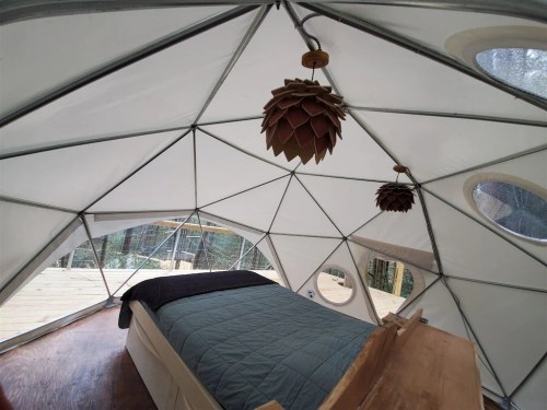 Interior of a geodesic dome with a bed, wooden accents, and unique pendant lights, surrounded by large windows.