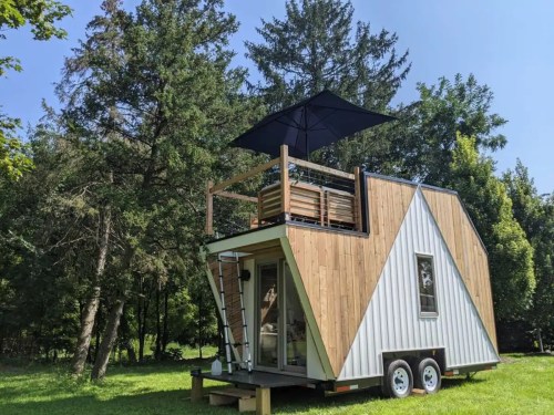 A modern tiny house on wheels with a rooftop deck and umbrella, surrounded by trees in a grassy area.