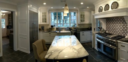 A modern kitchen featuring white cabinets, a large marble island, stainless steel appliances, and a patterned backsplash.