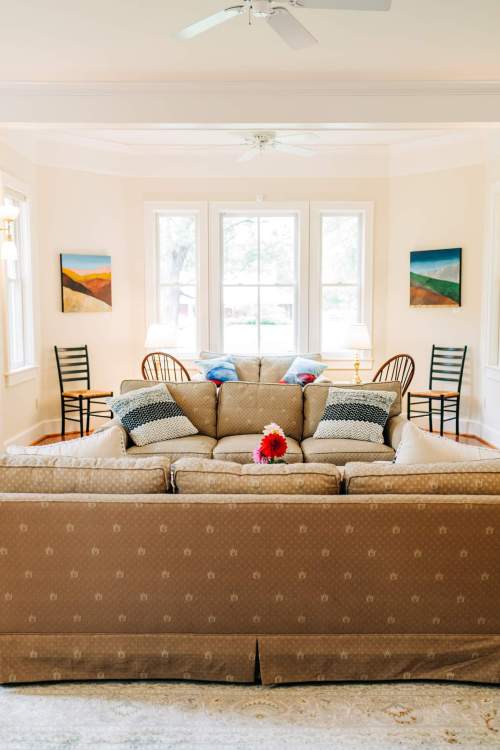 Cozy living room with a beige sofa, decorative pillows, and a view of a bright window with chairs and artwork.