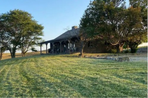 A rustic wooden house with a porch, surrounded by green grass and trees under a clear blue sky.
